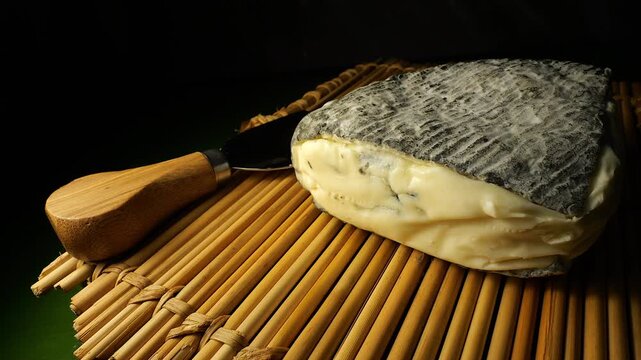 50p close up of Roche Baron cheese with ash rind displayed beside a cheese knife on a bamboo mat. Gourmet French dairy product in warm light