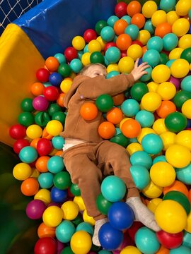 A joyful young boy in a cozy brown tracksuit having fun while lying and playing among numerous bright, multicolored plastic balls in a children's play area.