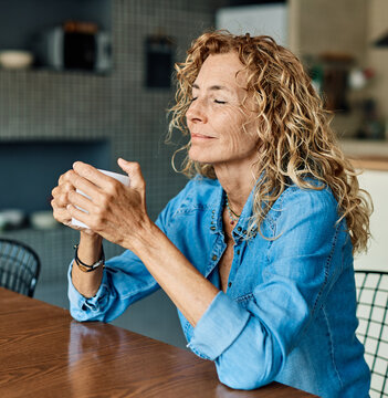 Portrait of a happy senior elderly woman holding a cup of coffee standing by window and relaxing at home