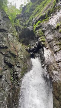 A beautiful vertical video of a crystal-clear waterfall flowing between rugged rock formations in a peaceful natural setting, this footage ideal for travel, adventure, relaxation, wellness, meditation
