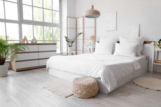 Interior of light room with white linen on bed, folding screen and plants