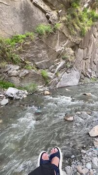 A relaxing vertical video featuring feet resting beside a flowing mountain stream surrounded by natural rocks and fresh water. Perfect for themes of travel, relaxation, mindfulness, outdoor adventure,