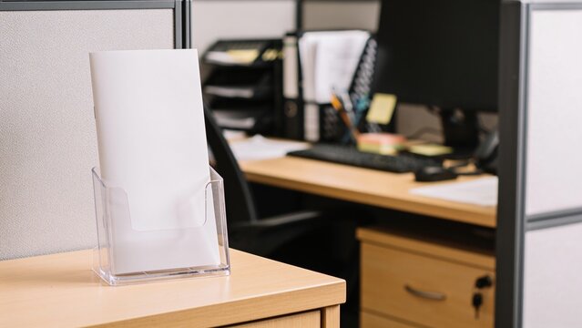 An office workspace with a clear acrylic brochure holder on a wooden desk. A computer and office supplies are visible in the background.