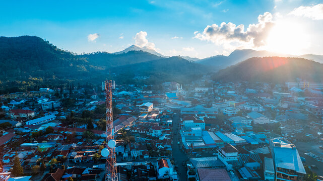 Aerial view of a telecommunications tower rising above the dense rooftops of the town, set against rolling mountains under the warm glow of a sunset in Bajawa, Nusa Tenggara Timur, Indonesia.