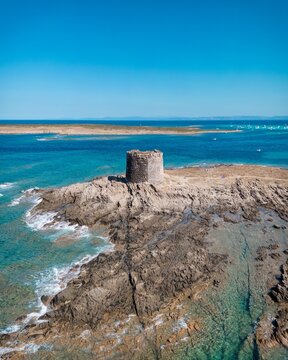 Aerial view of Spiaggia La Pelosa with its historic stone tower set on a rocky outcrop surrounded by clear turquoise Mediterranean waters in Sardegna, Italy.