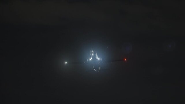 Airplane lifting into the night, bright landing lights and a glowing halo around the jet as it climbs head on after takeoff, red and green navigation lights on the wings, filmed in slow motion