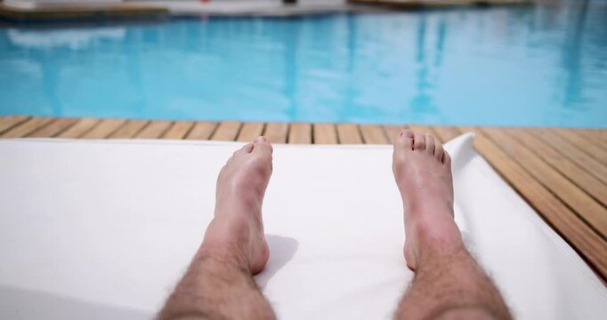 First person view relaxing beside a luxury swimming pool with feet stretched out on a white sun lounger beside clear blue water and wooden decking at a tropical holiday resort during summer travel