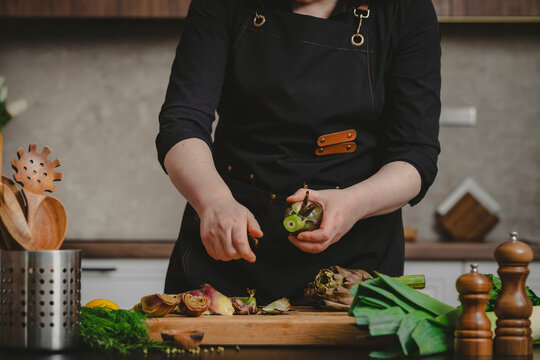 Chef hands trimming fresh green artichoke bottom with kitchen knife. Professional vegetable cleaning technique and organic culinary arts concept.