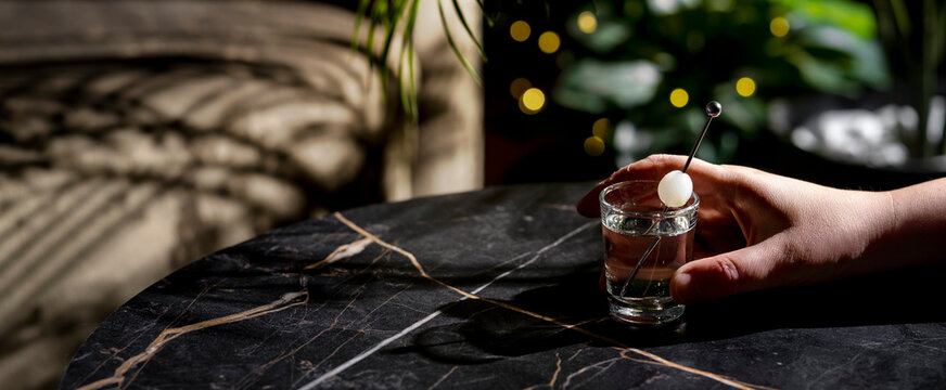 Hand reaching for tiniest tini savory martini cocktail glass on dark marble table at evening bar. Human touch in premium hospitality setting emphasizing nightlife experience and social drinking.