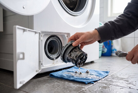 A person is cleaning a filter from a washing machine. Water spills onto the floor. The setting shows a laundry room with a washing machine and nearby items