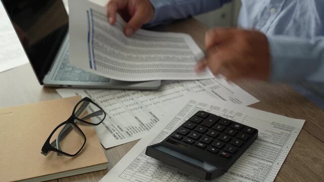 man use calculator while holding a financial document beside a laptop, showing budgeting, accounting, and report review workflow for corporate finance planning process.