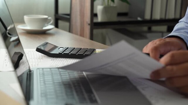 man use calculator while holding a financial document beside a laptop, showing budgeting, accounting, and report review workflow for corporate finance planning process.