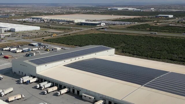 Reefer trucks line up at a cold storage port warehouse as a drone flies over vast rooftop solar panel arrays in daylight.