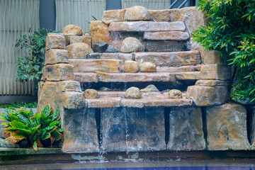 Stone waterfall cascades gently over tiered rock formations. Green foliage frames the natural water feature. Water pools softly at the base, reflecting calm surroundings © uniqephoto