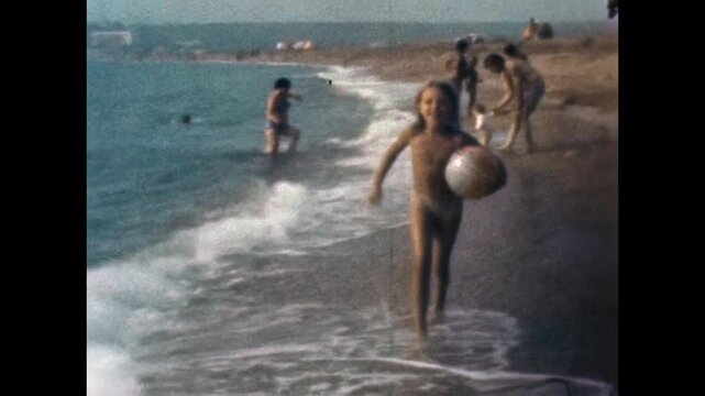Happy girl running along sandy seashore carrying ball during summer vacation. Joyful beach carefree childhood, outdoor play, active recreation by sea. Old film. Archival retro 1960s. Vintage archive
