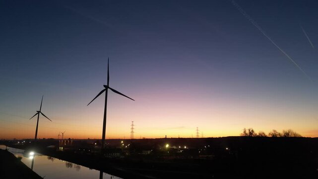 Aerial drone shot of wind turbines silhouetted against a beautiful sunrise, revealing a canal and industrial area.