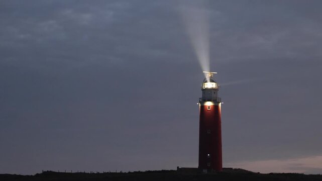 Red lighthouse on Texel Island shines a powerful rotating beam of light across the cloudy sky at dusk, symbolizing guidance and safety.