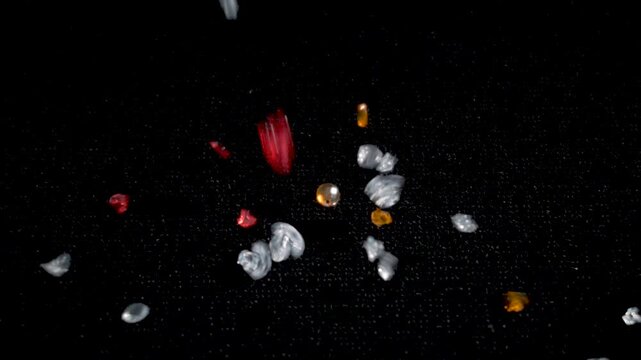 A close-up of small colorful beads and red flower-shaped ornaments falling against a black background captured in slow motion.