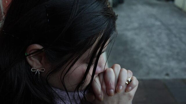 Touching close-up of a Latina girl in a purple fleece sweater, eyes closed, hands clasped in prayer outside on a chilly day. Soft natural light highlights her contemplative mood.