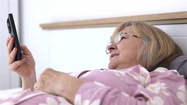 Senior woman in pajamas lying in bed and looking at smartphone at home, using mobile technology for communication or online browsing.