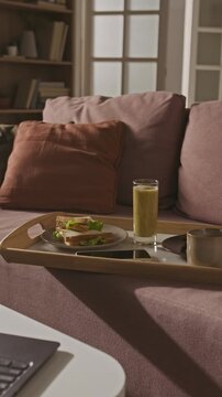 Vertical video of wooden tray holding healthy sandwich, green smoothie, smartphone, and coffee cup on sofa. Footage represents remote work lifestyle, balanced nutrition, and home office break