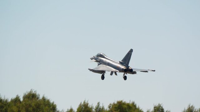 Combat aircraft on final approach with gear down and landing lights on in slow motion, military fighter jet seen head on against blue sky, warplane coming in to land