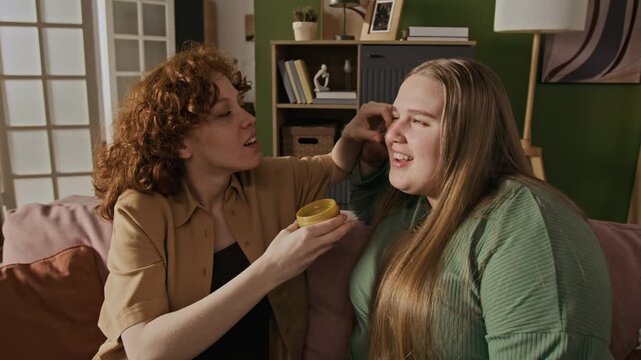 Young adult Caucasian pregnant woman applying cosmetic eye patches under eyes of her plus size female friend while sitting on sofa. Clip is ideal for skincare brand commercials and beauty tutorials