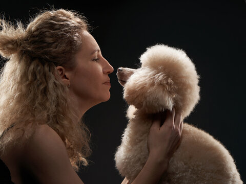 A young blonde woman lovingly lifts a white poodle close to her face. The intimate black studio background enhances the emotional connection between human and dog.