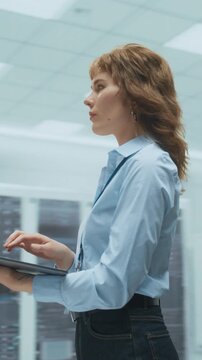 360 Degree Vertical Screen. Caucasian Woman Engineer Walks Through Modern Data Center Holding Laptop Computer, Reviewing Network Analytics, Configuring Servers, Overseeing Cloud Systems. Tracking Shot