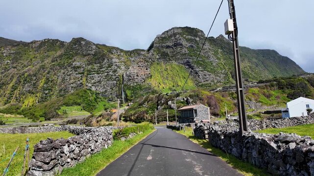 Walking along a quiet rural road surrounded by volcanic stone walls with Poco do Bacalhau waterfall and dramatic cliffs rising above on Flores Island Azores Portugal