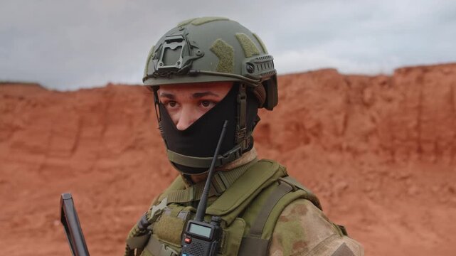 Soldier in tactical gear with radio and rifle on outdoor training range beside red clay berm under overcast sky, showing alert stance, protective helmet, dark face covering, and rugged field equipment