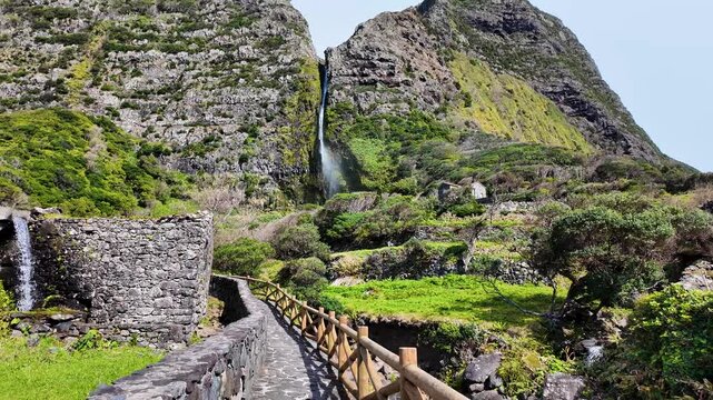 Walking along a stone pathway toward Poco do Bacalhau waterfall surrounded by lush vegetation volcanic cliffs and traditional landscapes on Flores Island Azores Portugal