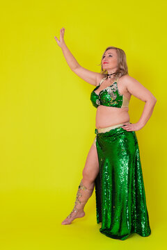 Sensuality mature woman belly dancer attired in dance stage outfit - green sequined bra top and maxi skirt with slit and she is not wearing any footwear, movement full length against yellow background