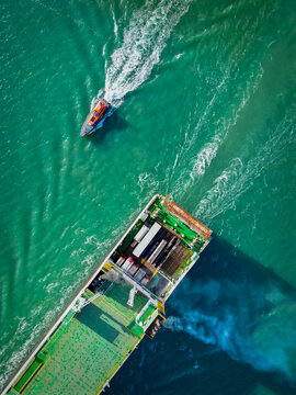 Aerial view of a large cargo ship navigating Dublin Harbour with a pilot boat nearby, leaving white wake trails in the green water of Dublin, County Dublin, Ireland.