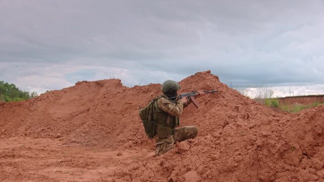 Soldier advancing with rifle across red dirt berm in sparse desert terrain, kneeling in tactical readiness under cloudy sky, with camouflage gear and focused combat posture