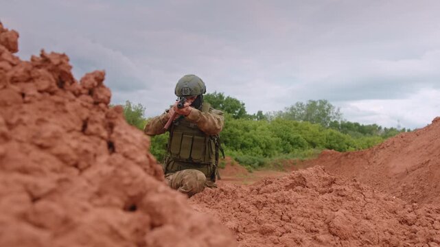Focused soldier in tactical gear kneeling behind red earth berm and aiming rifle across open training field under cloudy sky with calm natural light and clean background