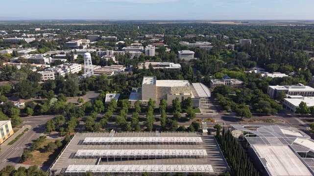 High-angle aerial perspective flying over the University of California, Davis, campus, showcasing modern architecture, parking structures with solar panels, and the surrounding verdant landscape