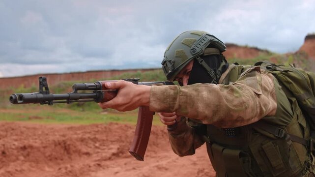 Soldier prone on desert training range aiming rifle under overcast sky, low-angle view with tactical gear, focused posture, red soil, cloudy light, clean copy space