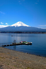 Fototapeta premium Snowy Fuji mountain lake pier boat blue sky sakura shoreline village landscape Snowy Fuji mountain above calm lake with pier and boat near shoreline village under blue sky with sakura season scenery