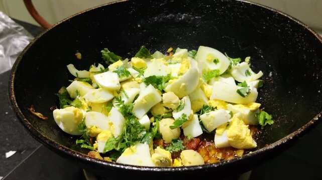 Close up of boiled egg pieces being added to rich Indian onion and tomato masala with fresh coriander in a traditional kadhai or pan  during home cooking.