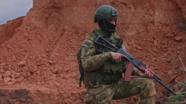 Kneeling soldier with rifle scanning left beside red earthen embankment, wearing olive camouflage gear and ballistic helmet during outdoor security training in soft daylight