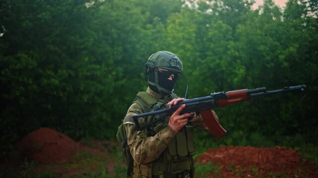 Camouflaged soldier adjusting rifle in wooded clearing with tactical vest, helmet, goggles and radio, alert stance during military training in green brown forest environment