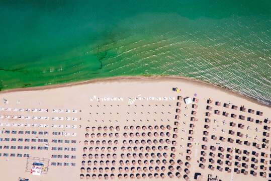 Aerial top down view of an organized sandy beach resort with rows of straw umbrellas and white sun loungers near the sea. Aerial top down view of rows of straw umbrellas and sun loungers on a sandy be