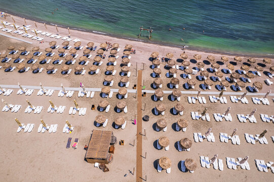 Aerial top down view of an organized sandy beach resort with rows of straw umbrellas and white sun loungers near the sea. Aerial top down view of rows of straw umbrellas and sun loungers on a sandy be