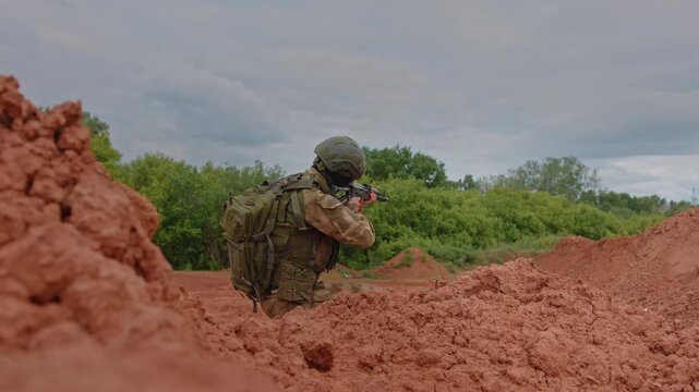Camouflaged soldier kneels behind red dirt berms with rifle raised, framed by blurred earth foreground, green tree line, and cloudy sky in tense outdoor training