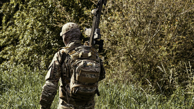 Camouflaged soldier in the middle of wilderness on a mission. Walking and aiming a AR-15 carbine rifle with a red collimator and a magnifier