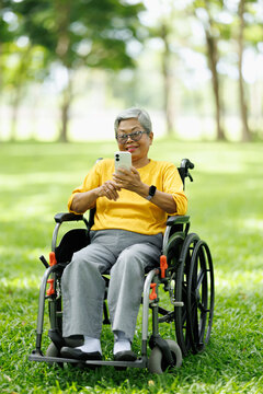 Senior asian woman in a wheelchair holding smartphone, smiling and messaging in a green park, representing active aging, independence, and digital connectivity