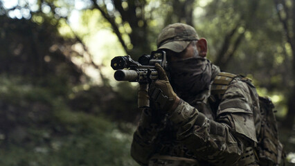 Fototapeta premium Camouflaged soldier in a dark forest on a recon mission. Hiding and aiming with a AR-15 carbine rifle with a laser collimator and a magnifier