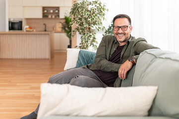 Fototapeta premium Mature man sitting comfortably on a modern couch, smiling cheerfully and looking directly at the camera, embodying a relaxed and happy lifestyle in his dwelling