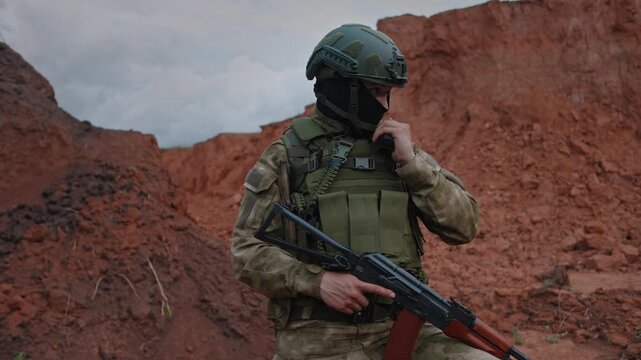 Camouflage soldier advances cautiously across red clay terrain beside eroded cliffs, holding rifle and handheld radio, with tense security patrol presence under cloudy natural light